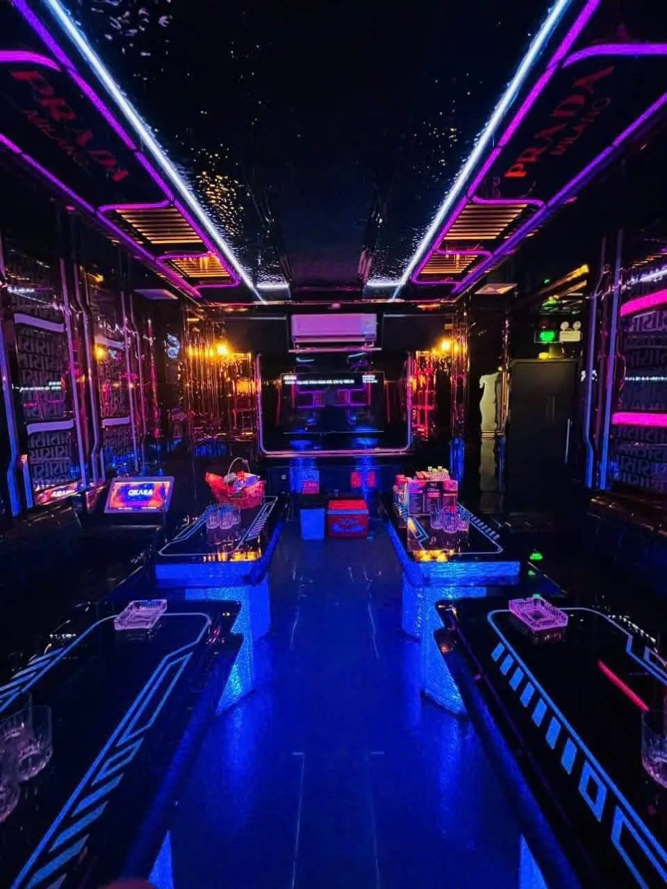 Phòng karaoke neon tím xanh có hai dãy bàn và lối đi giữa